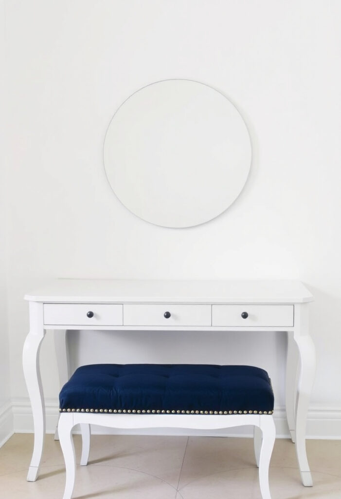a white vanity table with a round mirror above it. Underneath, a navy velvet upholstered bench.