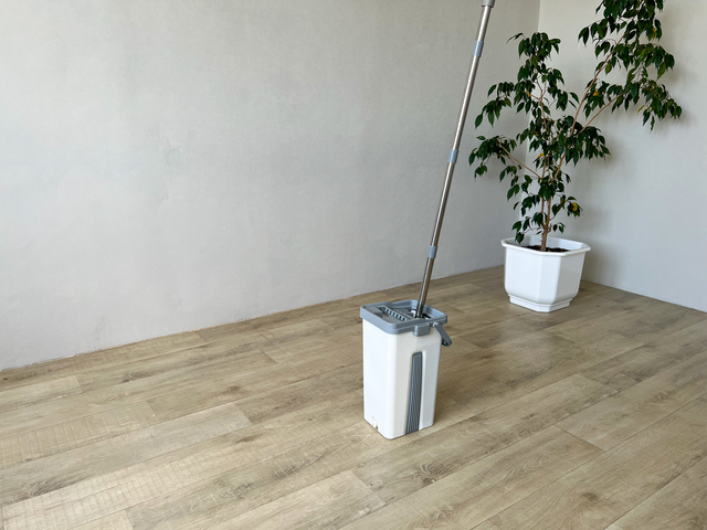 White plastic cleaning mop bucket with metal handle stands on laminate floor.